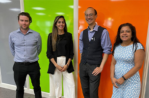 A group of 4 colleagues. Two are male and two are female. They are standing in a row against a green and orange wall. They are smiling and wearing smart clothes.